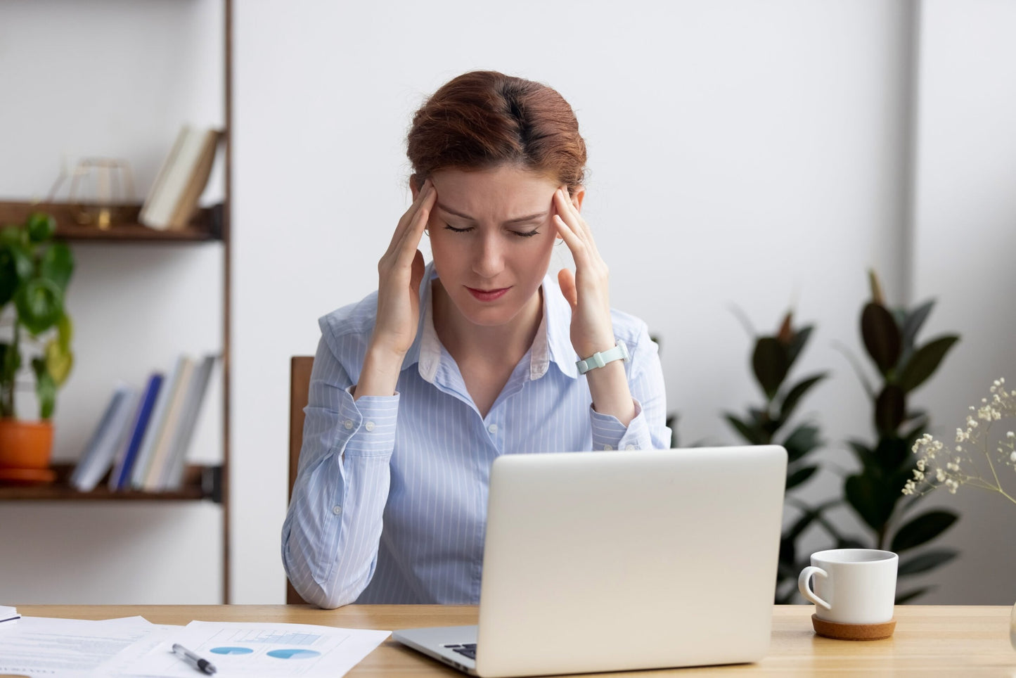 Photo of a woman sitting at her laptop with her eyes closed and her fingers on her temples, grimacing like she has a headache